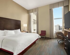 Bright hotel guest room with a king bed, white linens, dark wood headboard, tall windows with neutral curtains, bedside lamp, desk and chair, flat-screen TV, and city views.