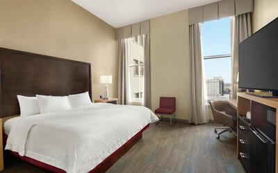 Bright hotel guest room with a king bed, white linens, dark wood headboard, tall windows with neutral curtains, bedside lamp, desk and chair, flat-screen TV, and city views.