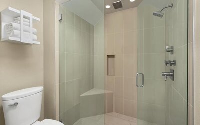 Modern hotel bathroom with a glass-enclosed walk-in shower, tiled walls, rainfall-style showerhead, built-in corner seat and wall niche, toilet, and open shelving with folded towels.
