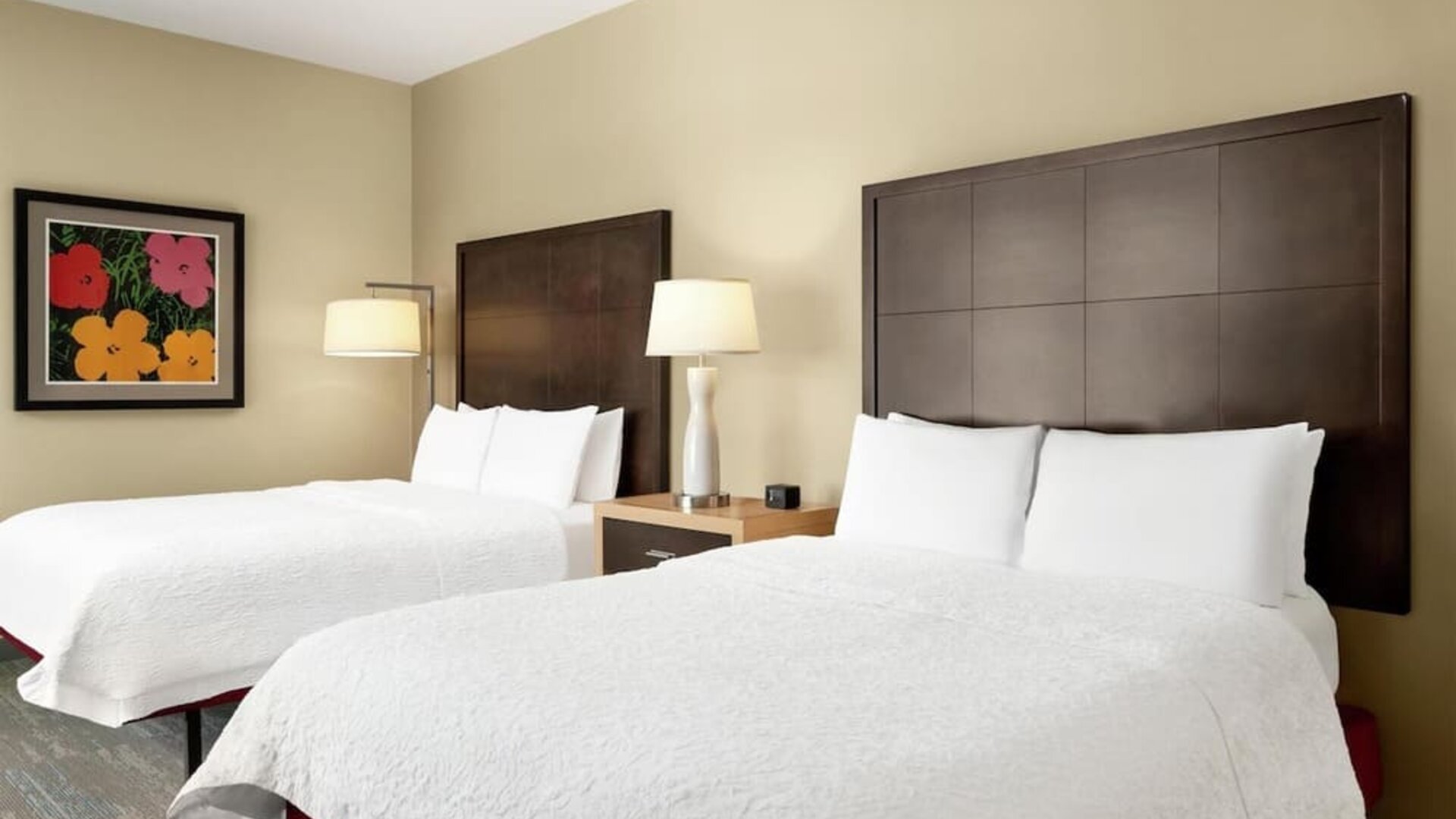 Hotel guest room with two beds dressed in white linens, tall dark wood headboards, shared nightstand with lamp, beige walls, and colorful floral artwork above one bed.