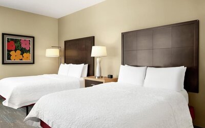 Hotel guest room with two beds dressed in white linens, tall dark wood headboards, shared nightstand with lamp, beige walls, and colorful floral artwork above one bed.