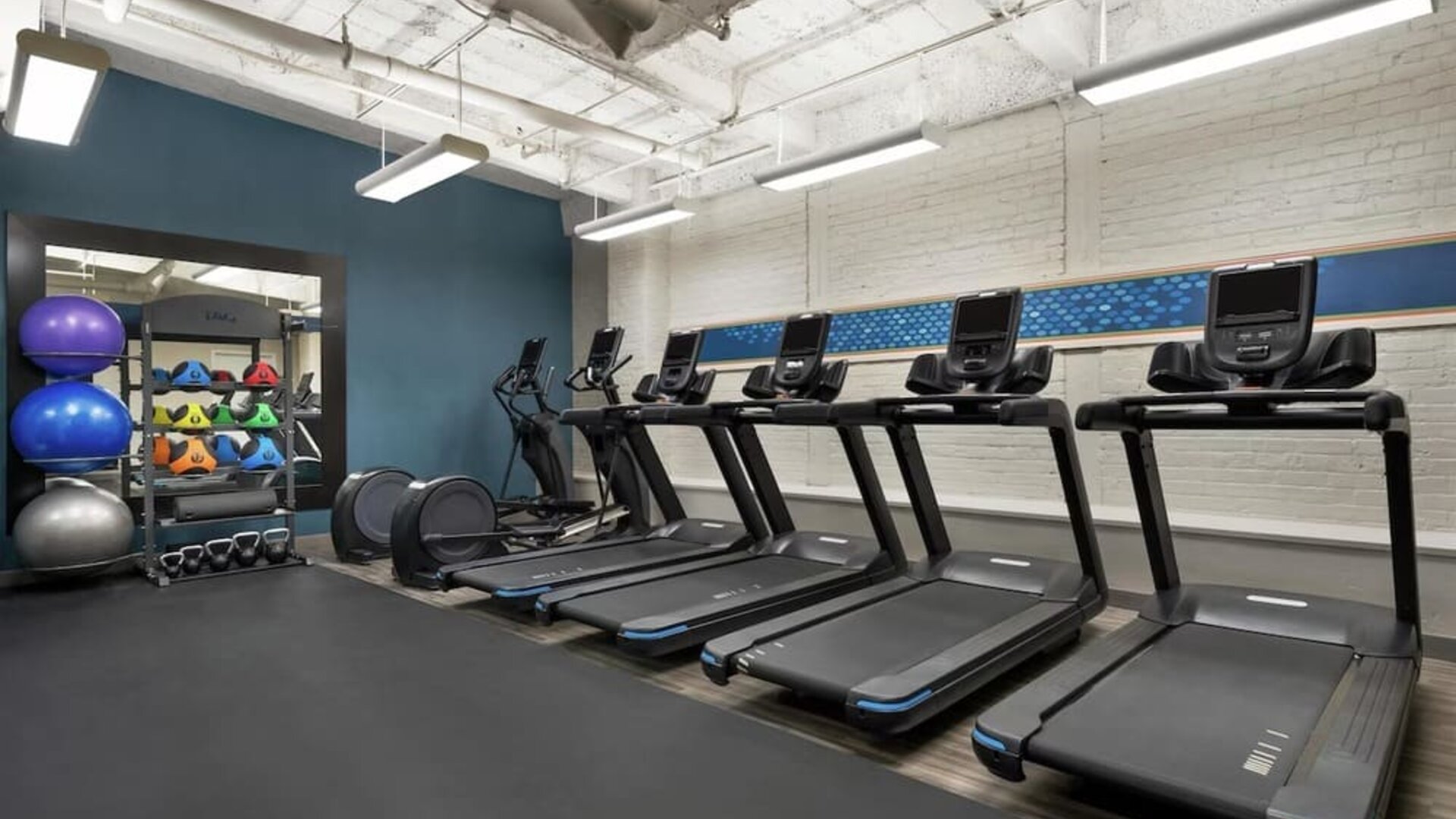Modern hotel fitness center with five treadmills, elliptical equipment, kettlebells, medicine balls, stability balls, white brick walls, and suspended rectangular lighting.