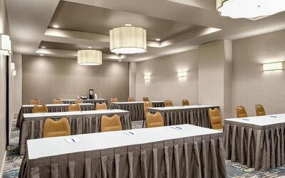 Hotel meeting room arranged classroom-style with long white tables, tan chairs, patterned carpet, large pendant lights, and notepads with pens at each place.