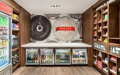 Hotel convenience shop with wooden shelving stocked with snacks, refrigerated cases containing bottled and canned drinks, and a central counter beneath a large “Treats” wall graphic.