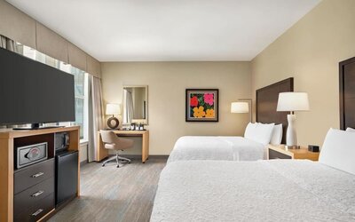Spacious hotel guest room with two beds dressed in white linens, dark wood headboards, bedside lamps, desk and chair, flat-screen TV, microwave, mini refrigerator, and colorful framed artwork.