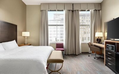 Spacious hotel room with a king bed, white linens, upholstered bench, desk and swivel chair, flat-screen TV, storage unit, tall windows, and a red accent chair overlooking the city.