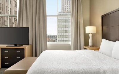 Hotel guest room with a king bed, white bedding, dark wood headboard, bedside lamp, flat-screen TV on a dresser, upholstered bench, and tall window overlooking surrounding city buildings.