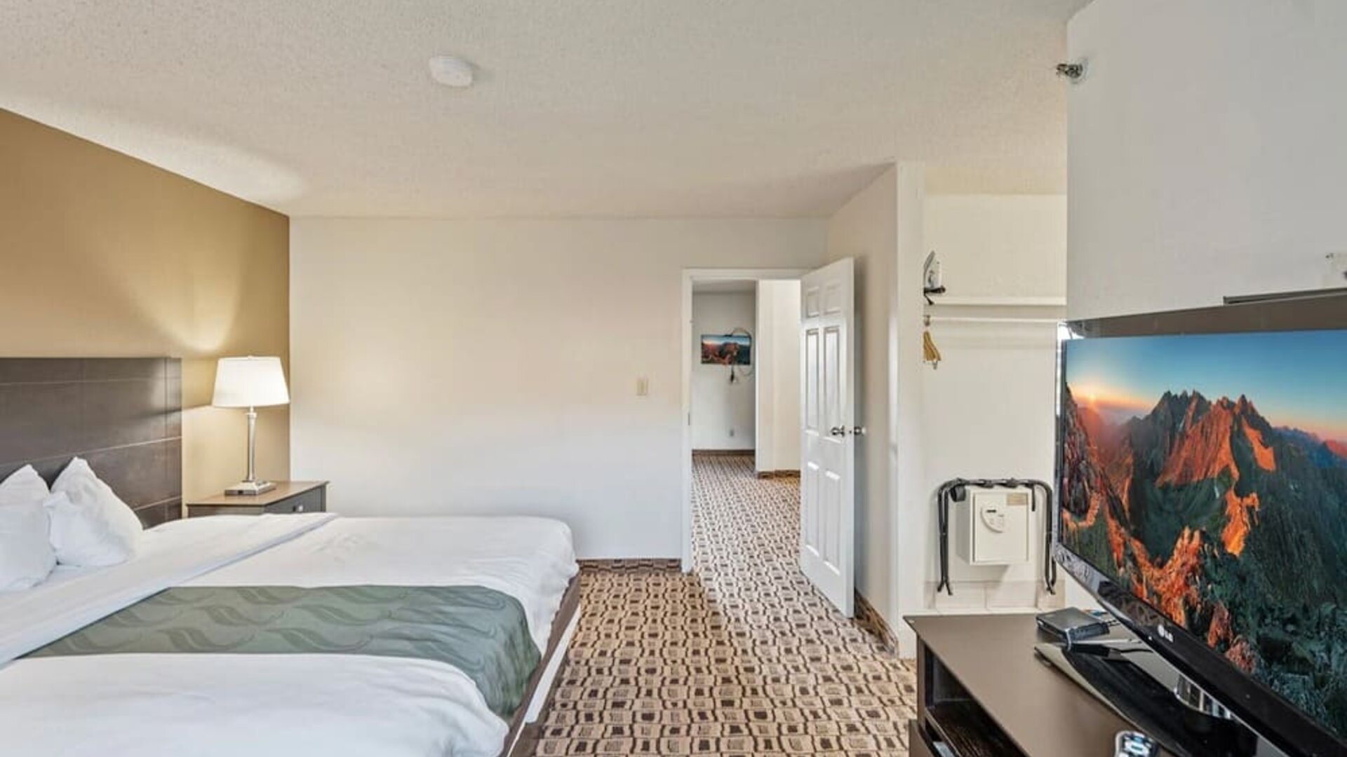 Spacious king guest room featuring a king bed, flat-screen television, storage space, and patterned carpeting at Coratel Plus Suites Wichita West Airport.
