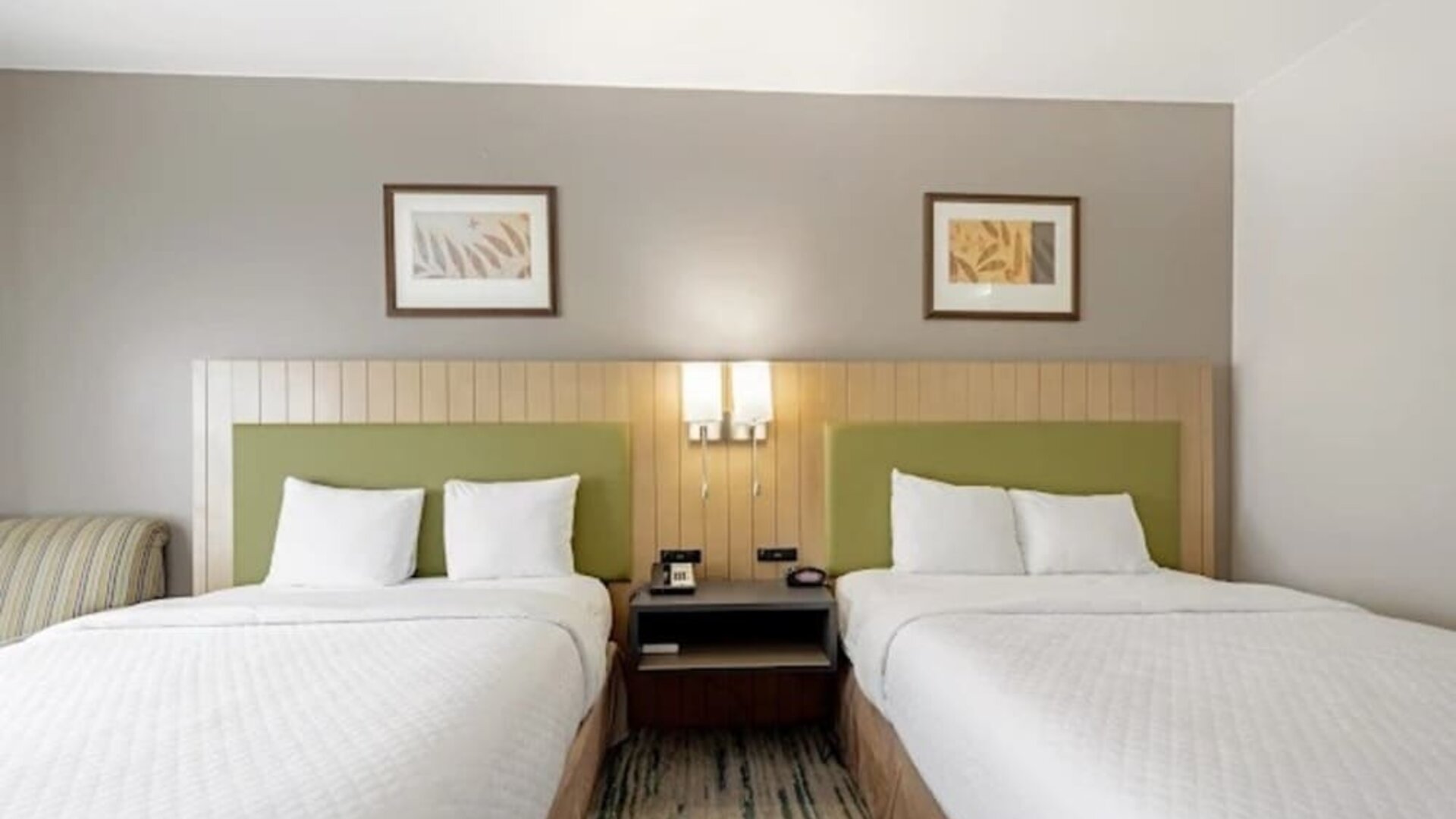 Guest room with two beds, white bedding, green upholstered headboards, shared nightstand, bedside lighting, and framed artwork at SureStay by Best Western Lewisville Dallas.