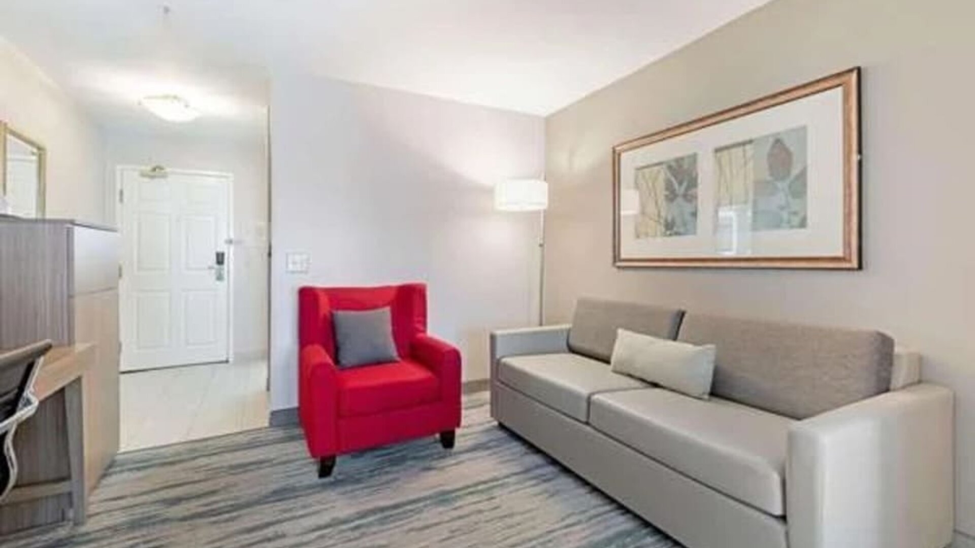 Guest suite seating area with sofa, red armchair, floor lamp, and framed artwork at SureStay by Best Western Lewisville Dallas.