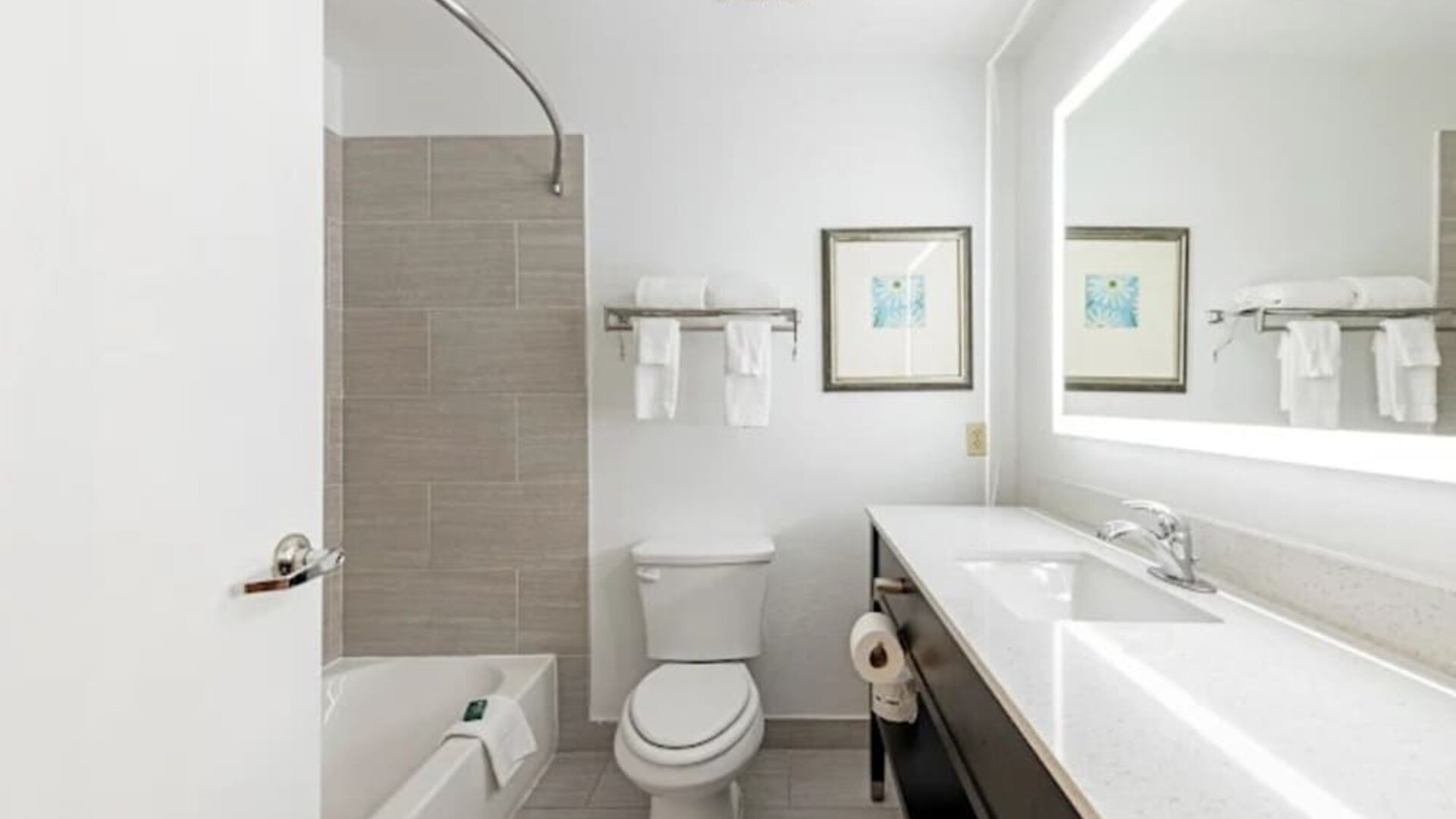 Guest bathroom with bathtub, tiled shower wall, toilet, long vanity, and illuminated mirror at SureStay by Best Western Lewisville Dallas.