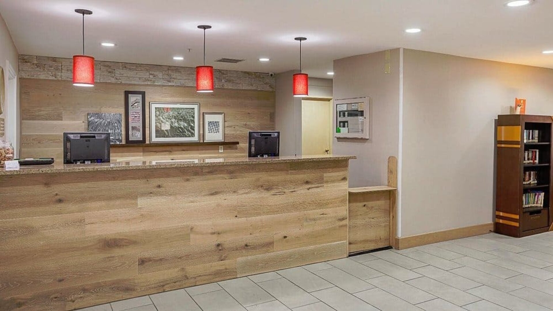 Reception area with wood-panelled front desk, red pendant lights, framed artwork, and bookcase at SureStay by Best Western Lewisville Dallas.