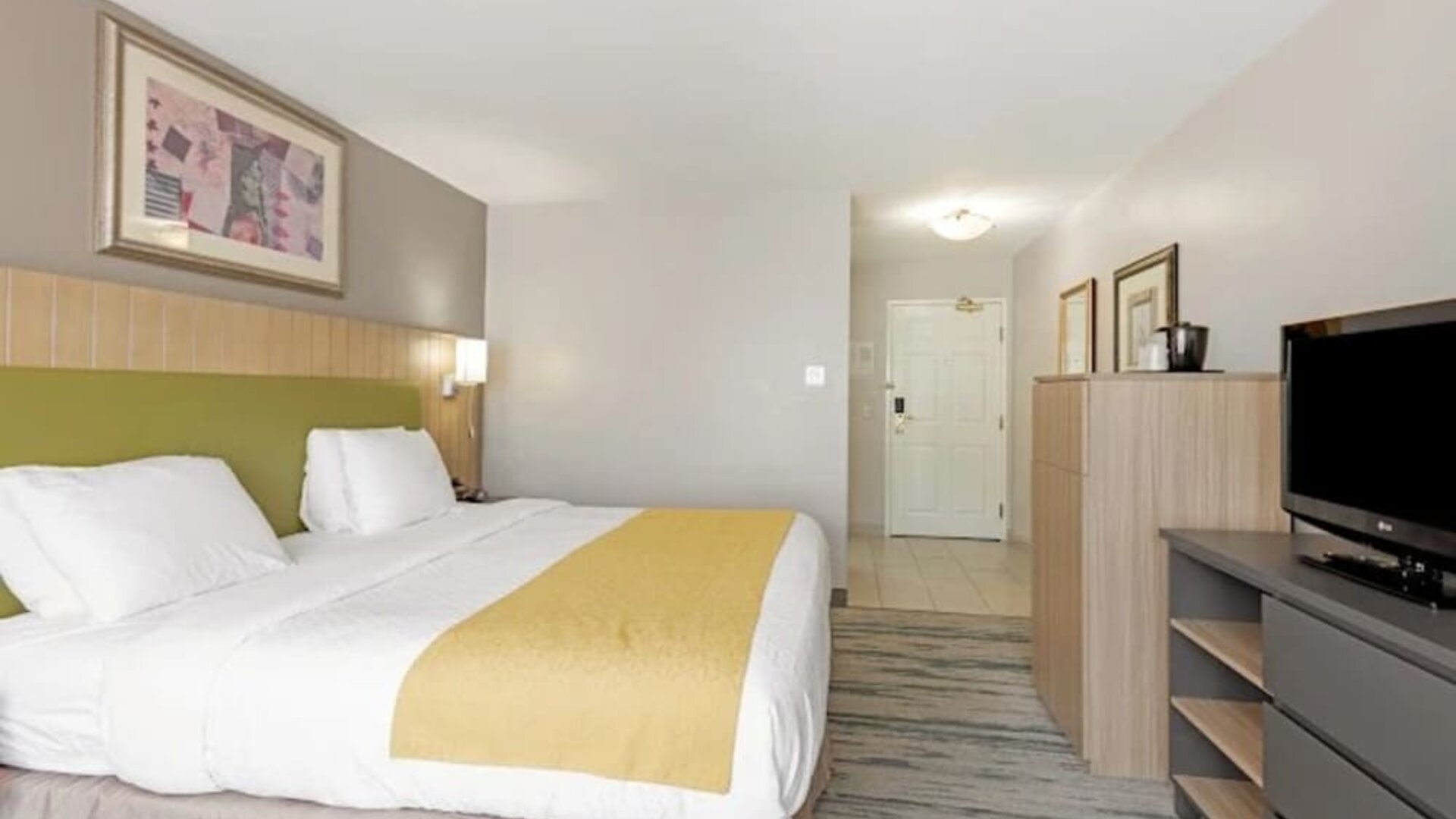King guest room with white bedding, yellow bed runner, green upholstered headboard, television, dresser, and storage cabinet at SureStay by Best Western Lewisville Dallas.