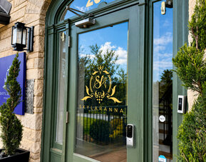 Main entrance to SKYLARANNA Resort & Spa, featuring glass doors with the resort logo, stone exterior, and decorative greenery.