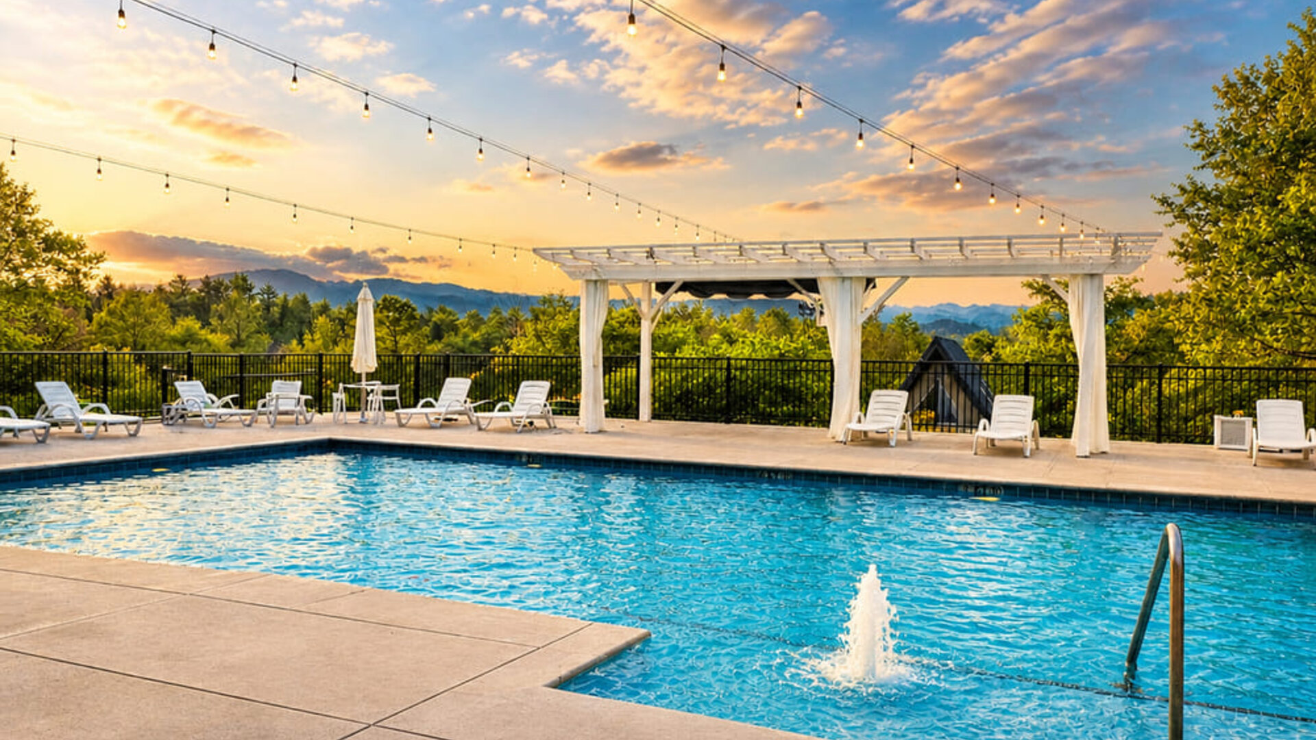 Outdoor swimming pool at sunset with sun loungers, a white pergola, string lights, and scenic mountain views at SKYLARANNA Resort & Spa.