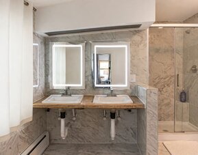 Guest bathroom at SKYLARANNA Resort & Spa, featuring double sinks, illuminated mirrors, marble-style finishes, and a glass-enclosed shower.