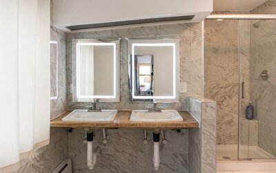Guest bathroom at SKYLARANNA Resort & Spa, featuring double sinks, illuminated mirrors, marble-style finishes, and a glass-enclosed shower.