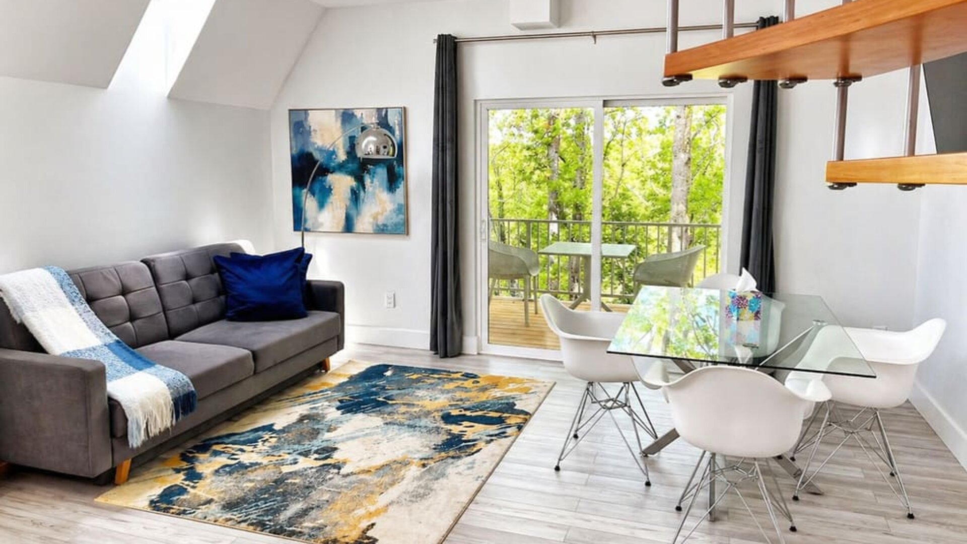 Open-plan cabin living and dining area at SKYLARANNA Resort & Spa, featuring a sofa, glass dining table, contemporary artwork, spiral staircase, and doors opening onto a private balcony.