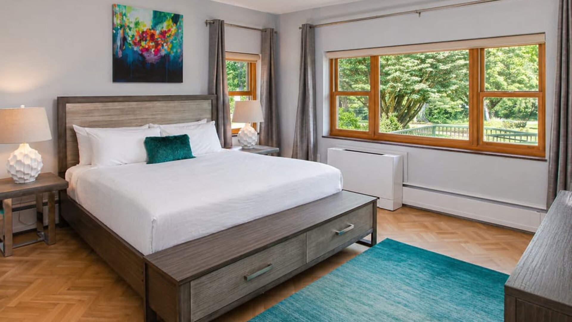 Guest room at SKYLARANNA Resort & Spa, featuring a king bed, contemporary furnishings, colourful artwork, large windows, and views of the surrounding greenery.