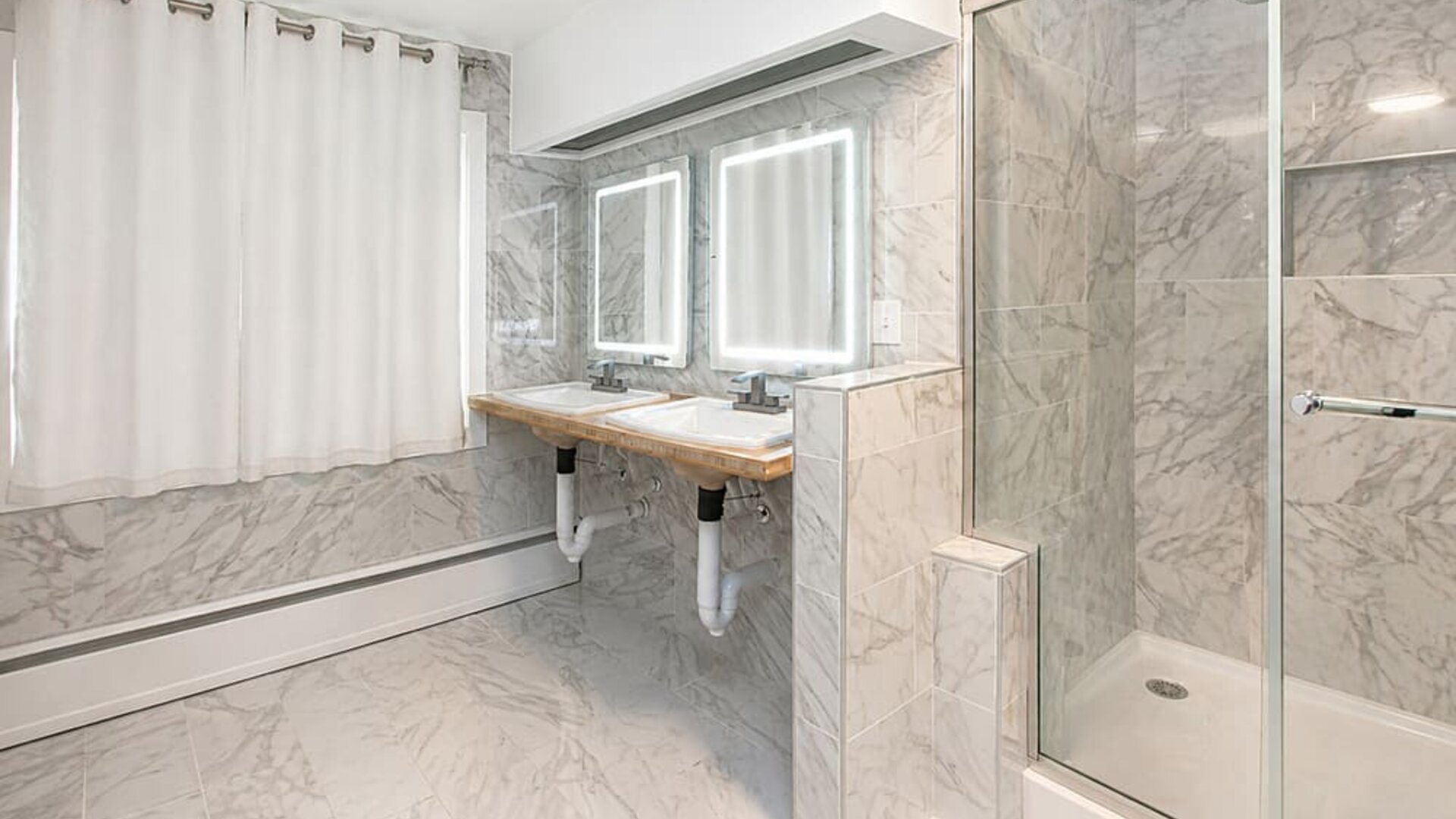 Spacious cabin bathroom at SKYLARANNA Resort & Spa, featuring double sinks, illuminated mirrors, marble-style finishes, and a glass-enclosed shower.