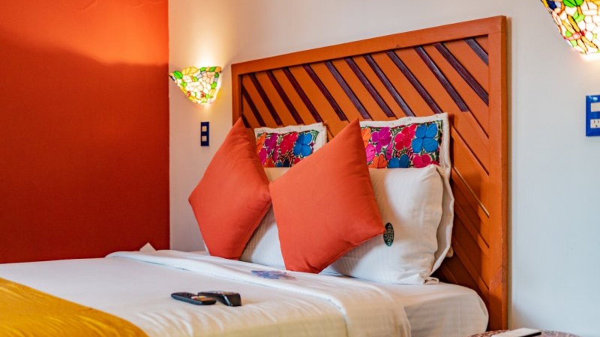 Colourful guest room featuring a comfortable bed, orange accent wall, decorative cushions, and stained-glass-style lighting at Casa Anzures Suites.