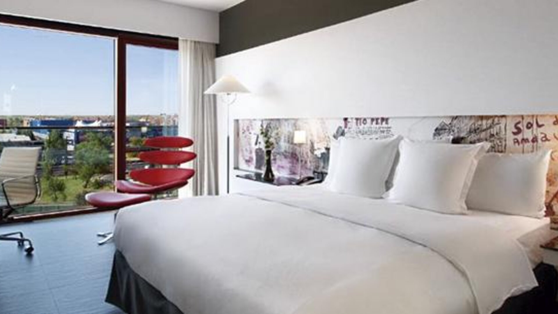Contemporary guest room featuring a large bed, dedicated workspace, colourful accent seating, and expansive windows with outdoor views at Hilton Madrid Aeropuerto.