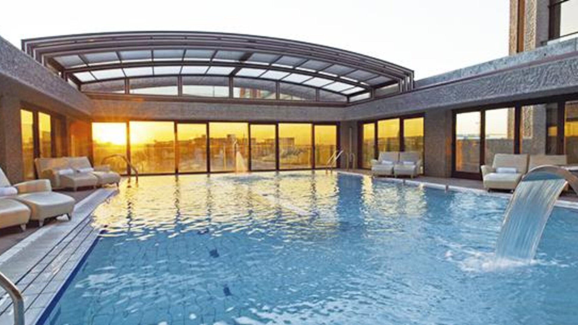 Spacious indoor swimming pool featuring a waterfall feature, poolside loungers, and expansive windows with sunset views at Hilton Madrid Aeropuerto.