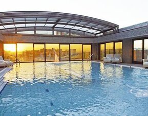 Spacious indoor swimming pool featuring a waterfall feature, poolside loungers, and expansive windows with sunset views at Hilton Madrid Aeropuerto.