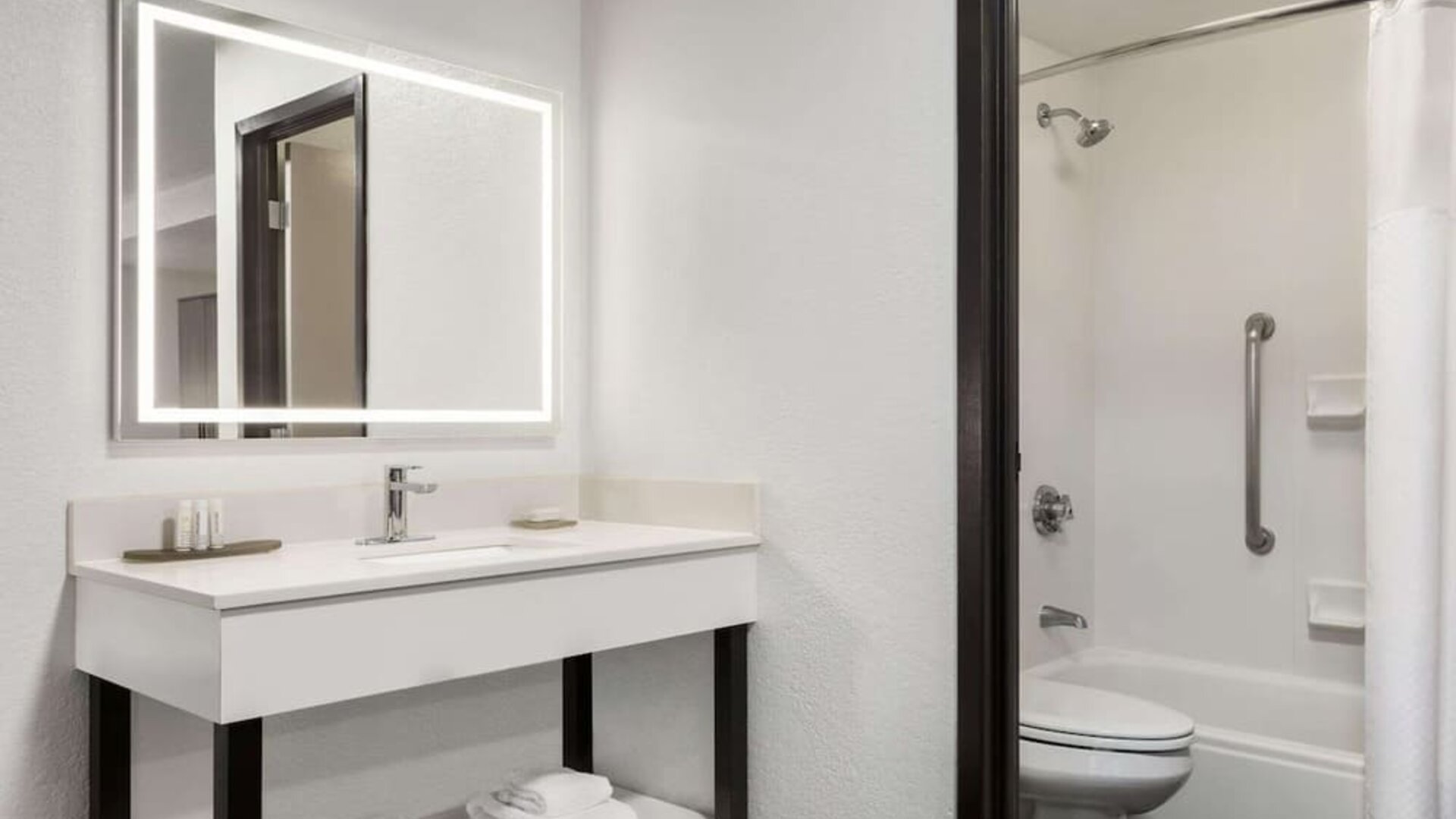 Bright guest bathroom featuring an illuminated mirror, spacious vanity, shower and bathtub combination, fresh towels, and accessibility grab bar at La Quinta Inn & Suites By Wyndham Cleveland Airport West.