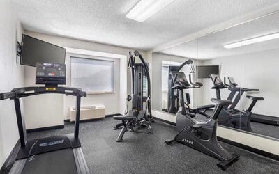 Compact hotel fitness center with treadmill, stationary bike, strength-training machine, wall-mounted TV, and mirrored wall.