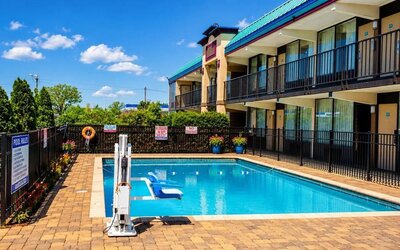Rectangular outdoor hotel swimming pool surrounded by a brick-paved deck and black safety fencing, with an accessible pool lift and two-story guest room building alongside.