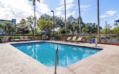 Outdoor hotel swimming pool surrounded by a concrete deck, lounge chairs, tables with umbrellas, palm trees, safety fencing, and an accessible pool lift.