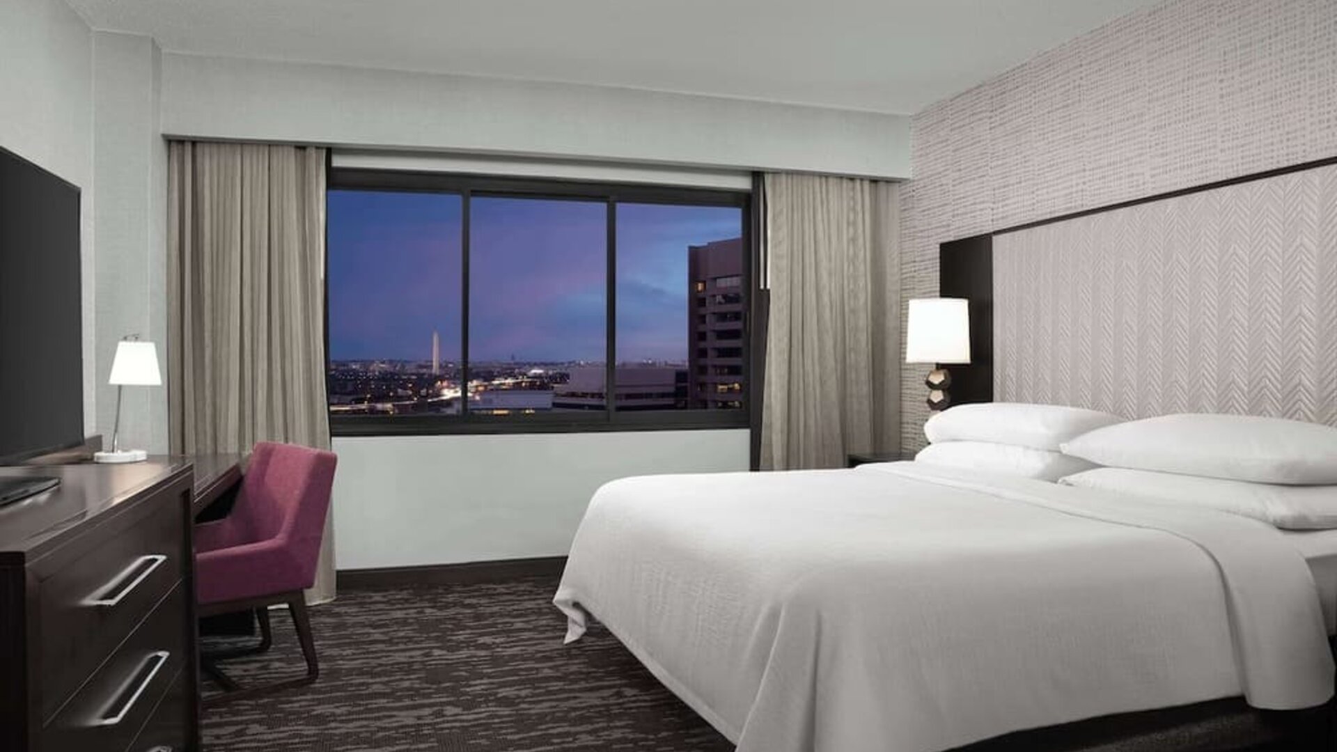 Comfortable king guest room featuring a large bed, dedicated workspace, television, and expansive window with city views at Embassy Suites by Hilton Crystal City National Airport.