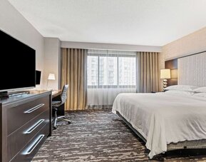 Bright king guest room featuring a comfortable bed, workspace, television, and ample natural light from a large window at Embassy Suites by Hilton Crystal City National Airport.