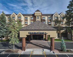 Daytime exterior view of the multi-story DoubleTree by Hilton hotel, featuring a covered entrance with hotel signage, beige facade, peaked rooflines, mature evergreen trees, and a paved driveway.
