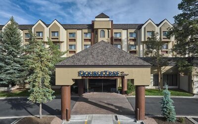 Daytime exterior view of the multi-story DoubleTree by Hilton hotel, featuring a covered entrance with hotel signage, beige facade, peaked rooflines, mature evergreen trees, and a paved driveway.