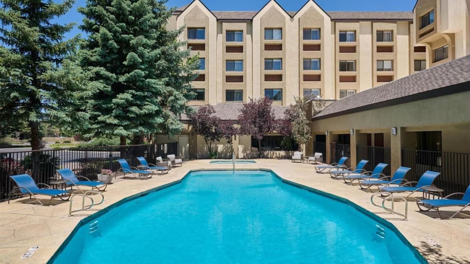 Large rectangular outdoor swimming pool surrounded by a sunny patio with blue lounge chairs, tables, mature evergreen trees, and the multi-story hotel building in the background.