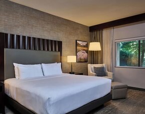 Modern hotel room with a king bed dressed in crisp white linens, tall dark headboard, bedside lamps, upholstered armchair and ottoman, framed artwork, and a large window overlooking green trees.