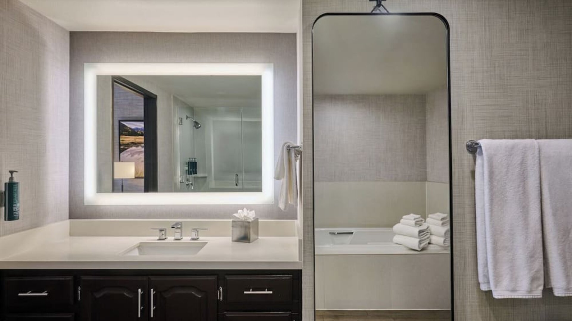 Modern hotel bathroom with a wide illuminated vanity mirror, white countertop and sink, dark wood cabinetry, towel rack, and a bathtub visible through a tall wall mirror.