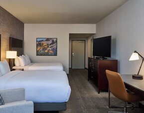 Spacious hotel guest room with two beds in white linens, dark headboards, an upholstered armchair, flat-screen television on a dresser, and a desk with lamp, chair, and round wall mirror.
