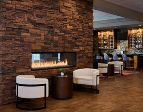 Cozy hotel lounge featuring a large stacked-stone fireplace with visible flames, two cream armchairs and a side table, with a warmly lit bar and counter seating in the background.