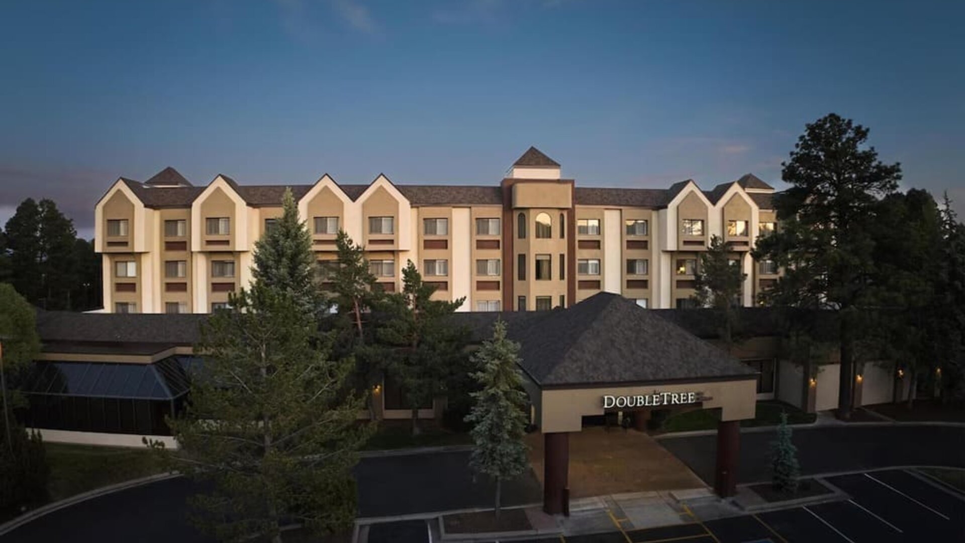 Elevated dusk view of the DoubleTree hotel exterior with illuminated entrance signage, multi-story beige facade, surrounding pine trees, dark parking area, and accessible parking spaces near the entrance.