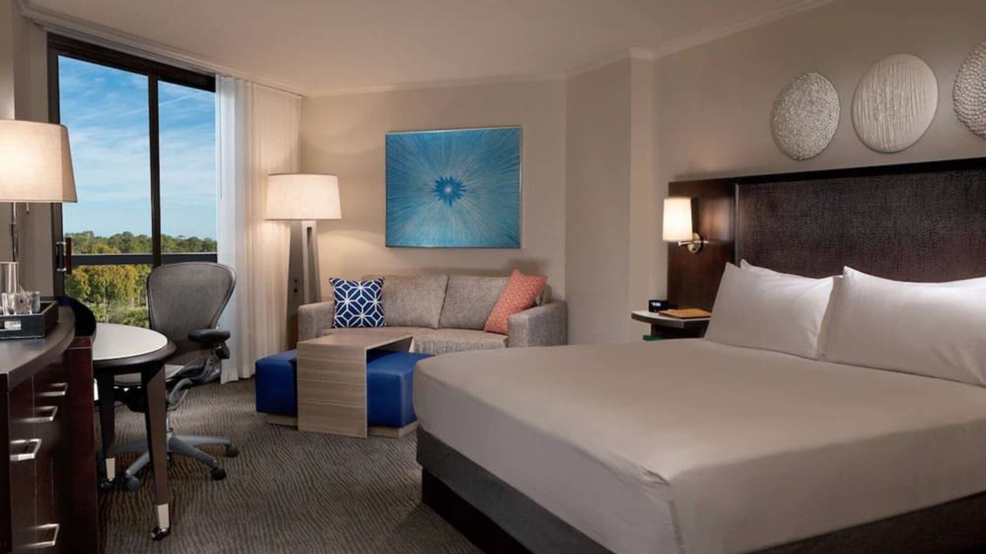 Spacious hotel room with a king bed, white linens, dark upholstered headboard, sofa with colorful accent pillows, work desk and chair, blue artwork, and a large window overlooking trees.