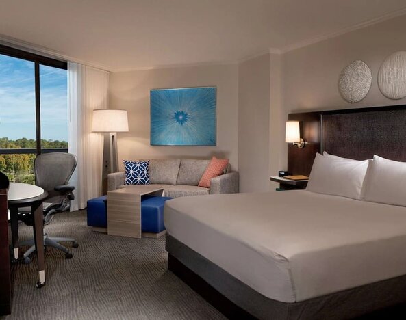 Spacious hotel room with a king bed, white linens, dark upholstered headboard, sofa with colorful accent pillows, work desk and chair, blue artwork, and a large window overlooking trees.