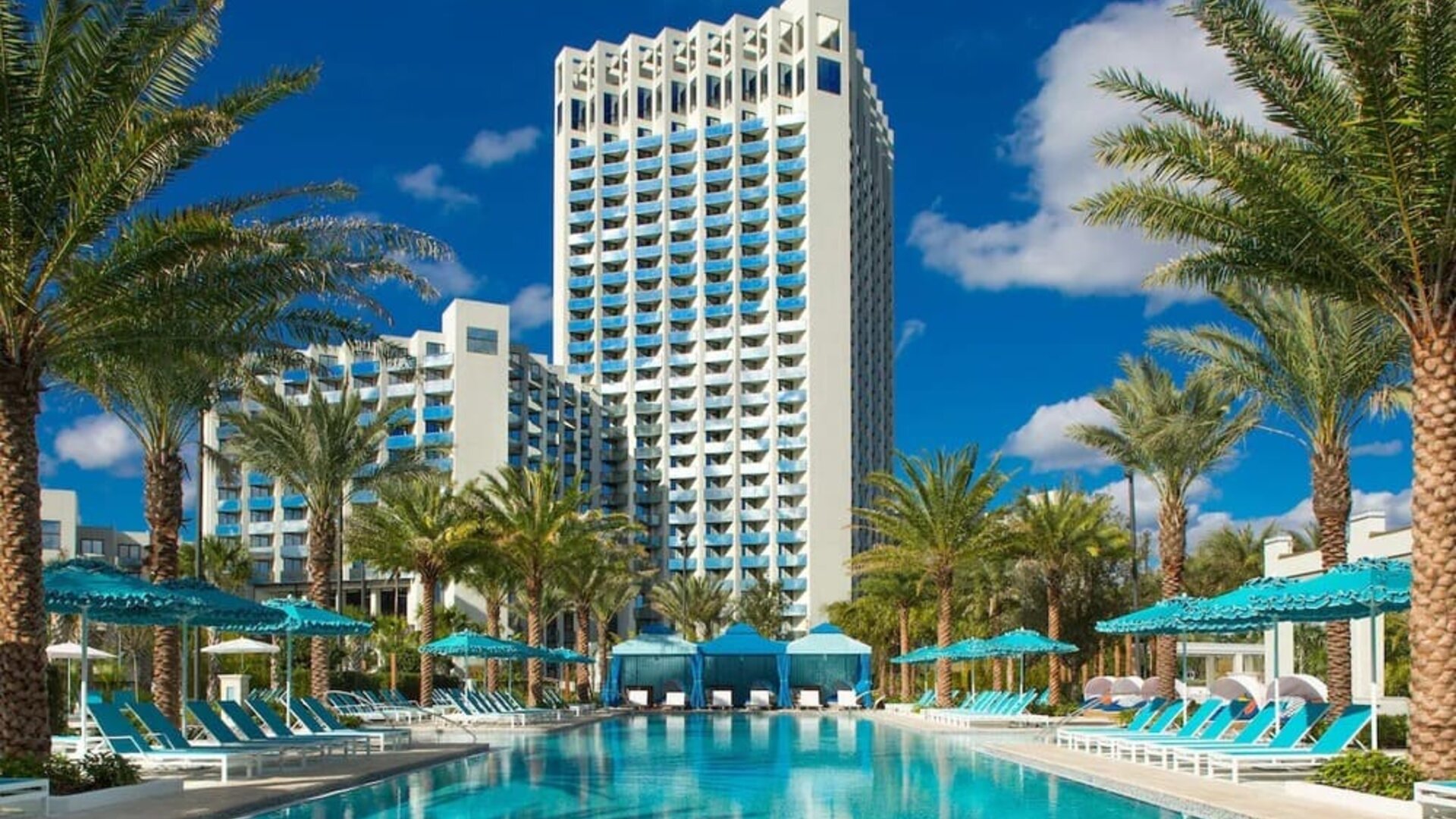 Palm-lined resort swimming pool with turquoise water, rows of blue lounge chairs and umbrellas, private cabanas, and a tall white hotel tower rising in the background.