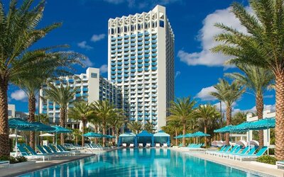 Palm-lined resort swimming pool with turquoise water, rows of blue lounge chairs and umbrellas, private cabanas, and a tall white hotel tower rising in the background.