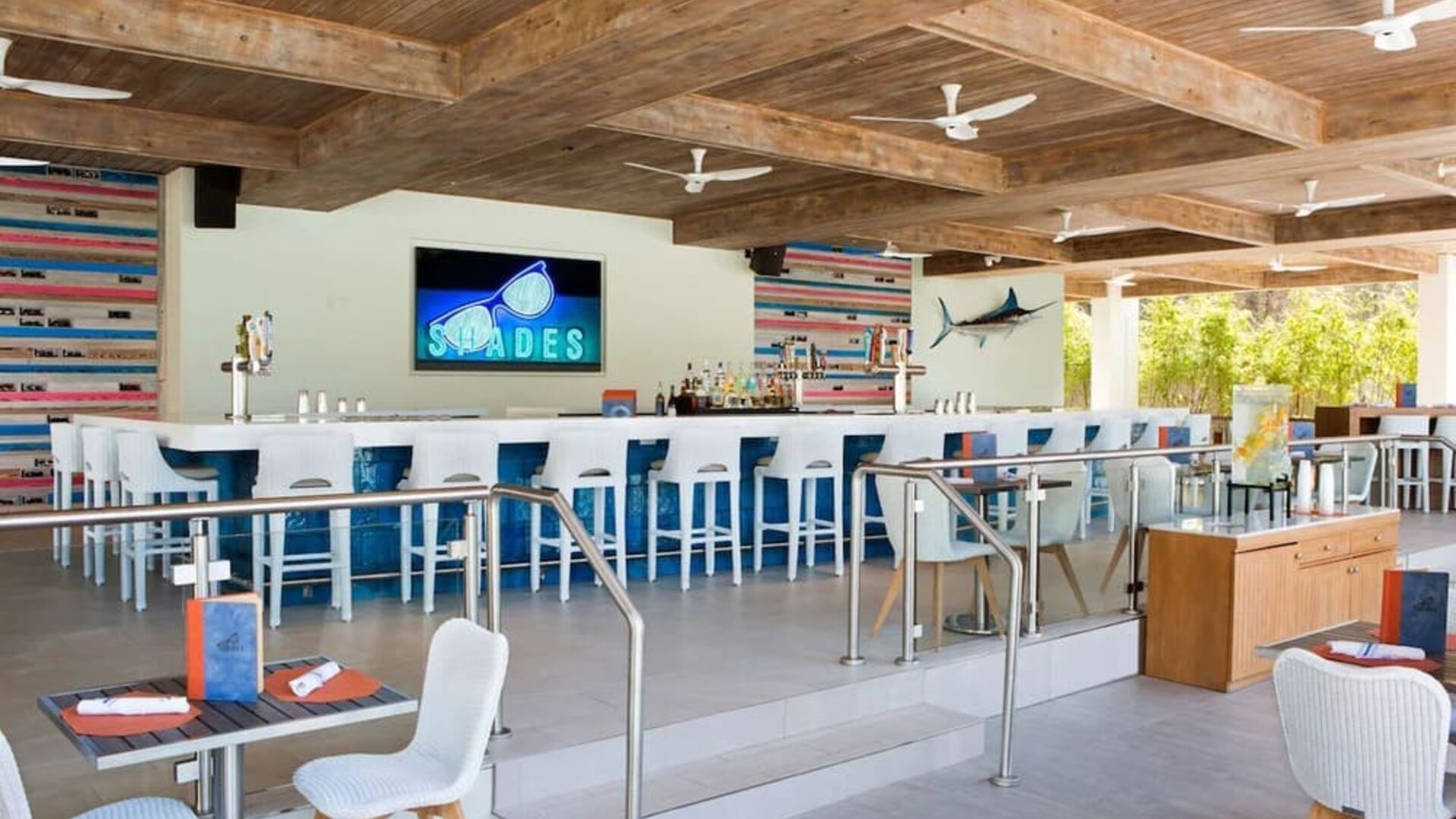 Open-air resort bar with long white counter, blue-and-white stools, beverage taps, colorful coastal decor, dining tables, exposed wood ceiling beams, and multiple ceiling fans.