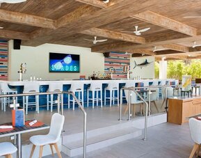 Open-air resort bar with long white counter, blue-and-white stools, beverage taps, colorful coastal decor, dining tables, exposed wood ceiling beams, and multiple ceiling fans.