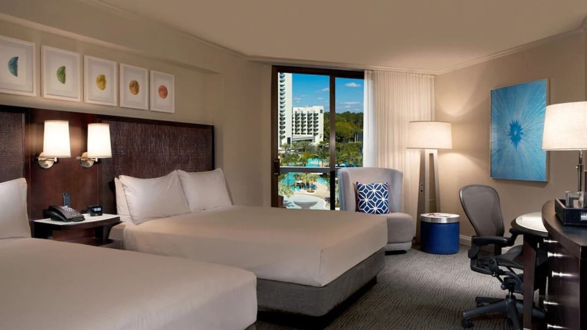 Spacious hotel room with two beds dressed in white linens, dark headboard with reading lights, armchair, work desk and ergonomic chair, blue accent decor, and a window overlooking the resort pool area.