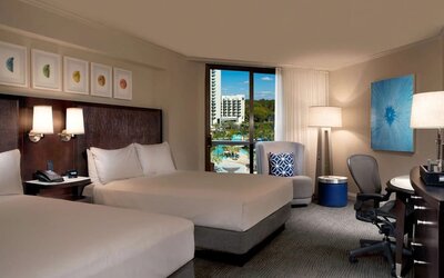 Spacious hotel room with two beds dressed in white linens, dark headboard with reading lights, armchair, work desk and ergonomic chair, blue accent decor, and a window overlooking the resort pool area.